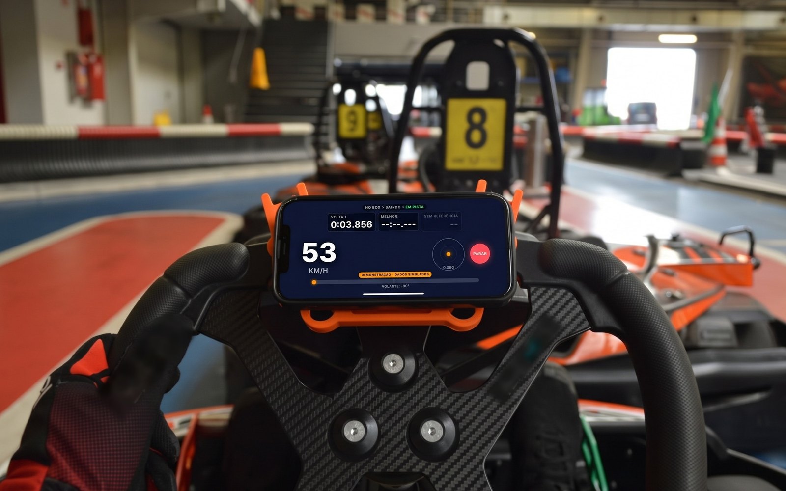 iPhone preso no volante de um kart, com a tela do Kart Coach mostrando velocidade e tempo de volta, karts alinhados ao fundo.